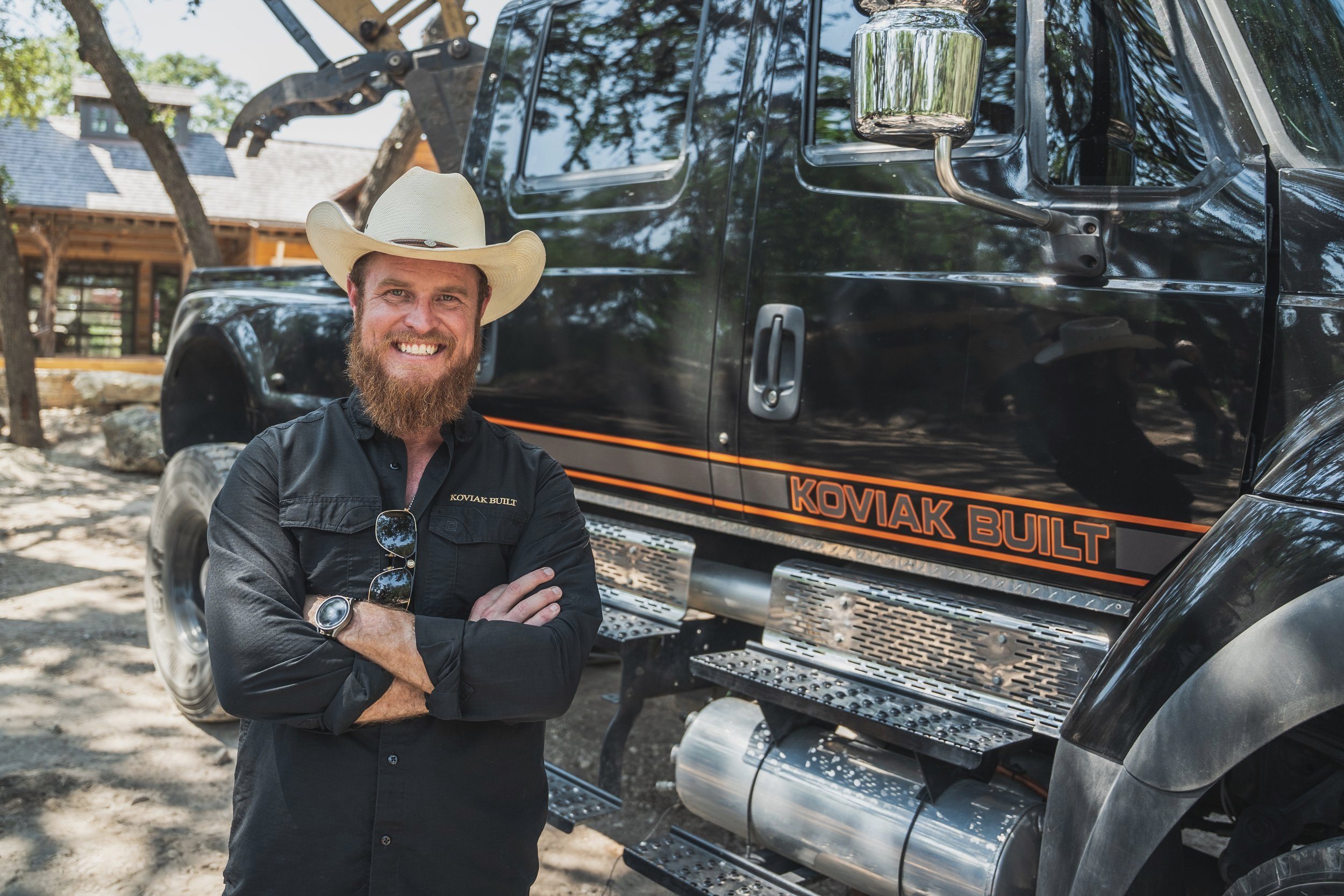 Photo of Joshua Koviak standing in front of a large truck wearing a black Koviak Built shirt and a cowboy hat