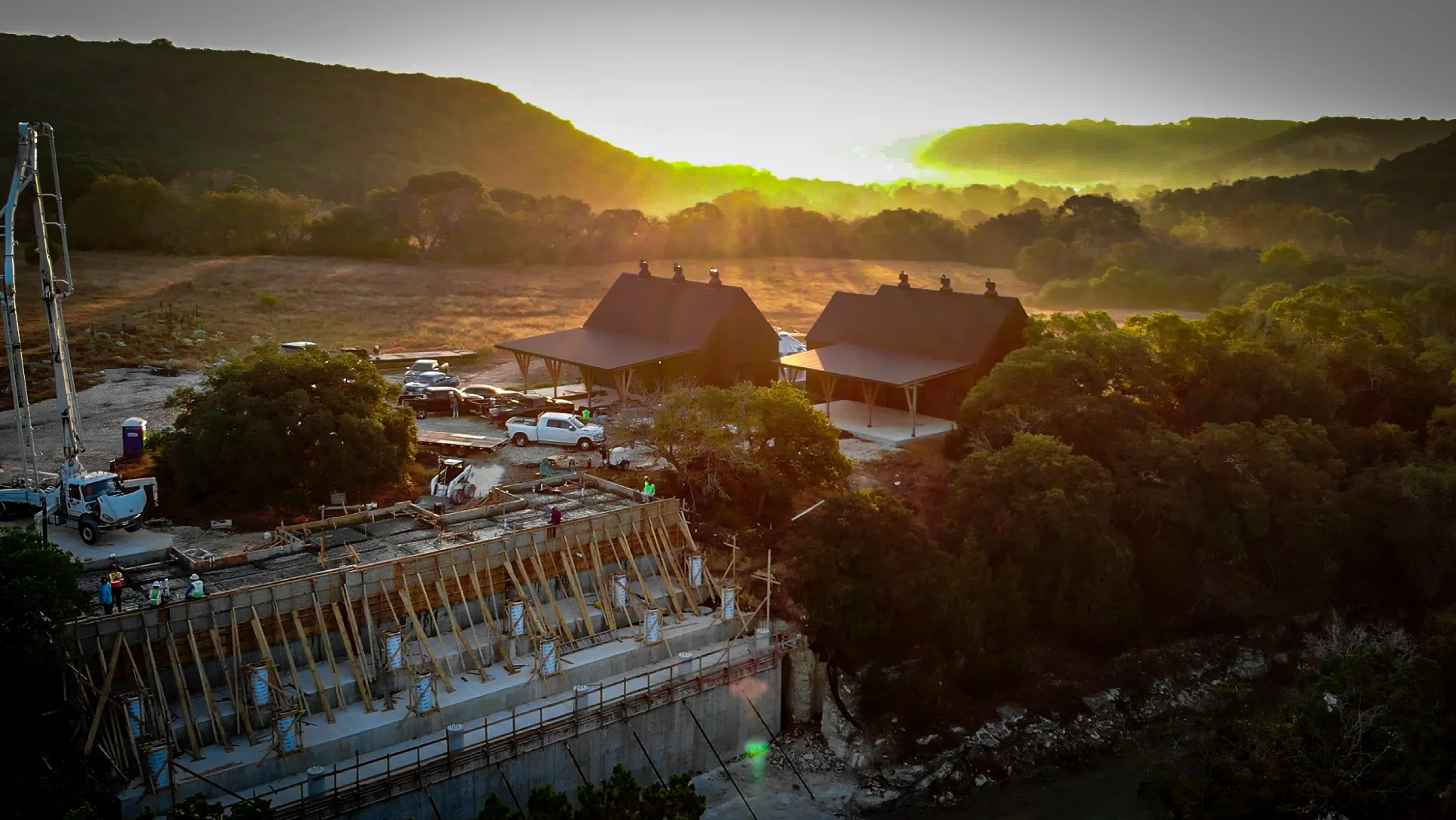 Arroyo Verde aerial construction view at sunset with foundation work, neighboring roofs, and Hill Country hills